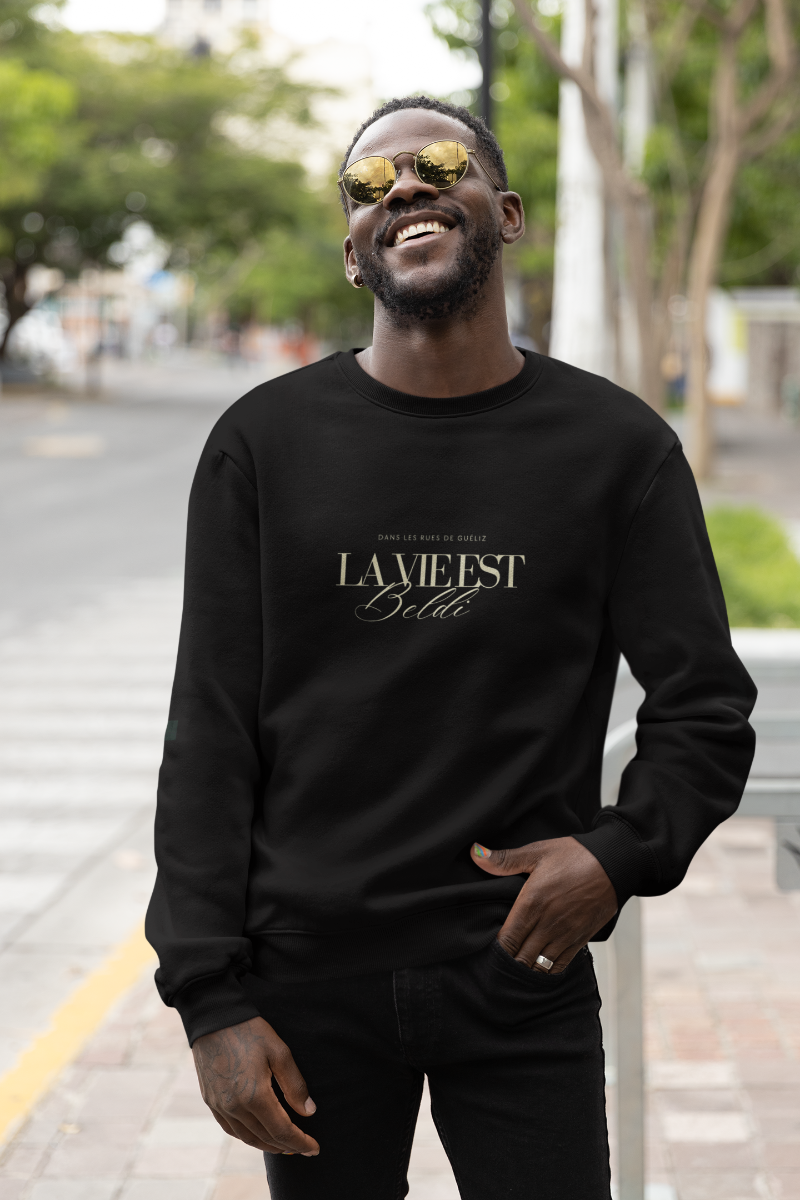  Man smiling in sunglasses and a black “La Vie Est Beldi” sweatshirt, walking on a city sidewalk, embracing Moroccan-inspired street style.