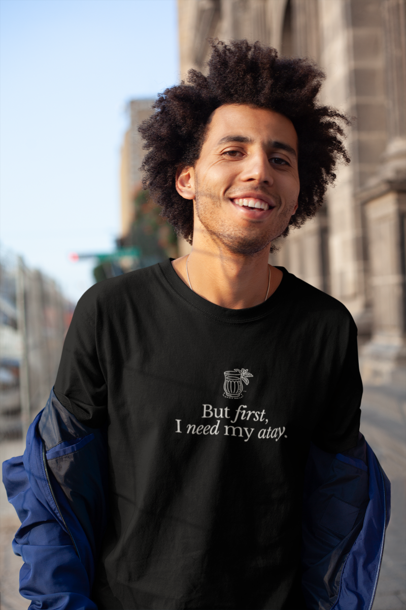 Smiling man wearing a 'But First, I Need My Atay' t-shirt, enjoying a sunny day. Trendy Moroccan-inspired tea lover shirt for a relaxed and casual look