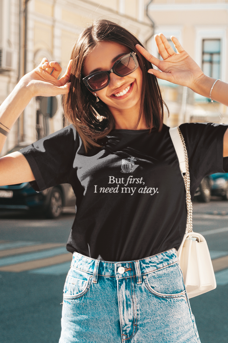 Smiling woman enjoying a sunny day while wearing a 'But First, I Need My Atay' t-shirt. Stylish Moroccan-inspired tea lover shirt, perfect for casual and cozy vibes