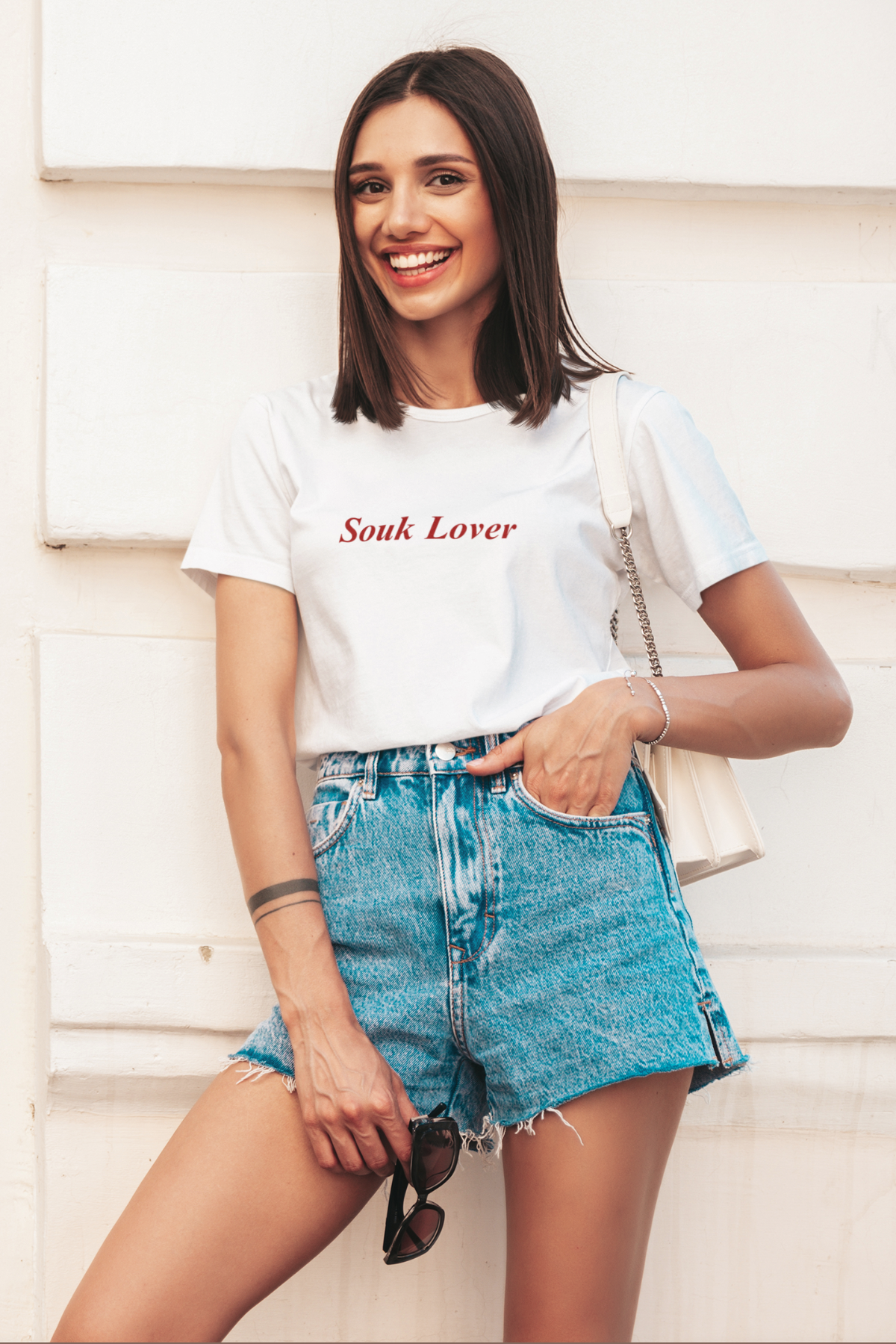 Woman in off-white ‘Souk Lover’ T-shirt with red text, holding sunglasses and smiling